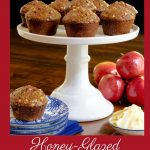 Vertical photo of a batch of Honey-Glazed Apple Bran Muffins on a white pedestal plate surrounded by apples and a dish of butter.