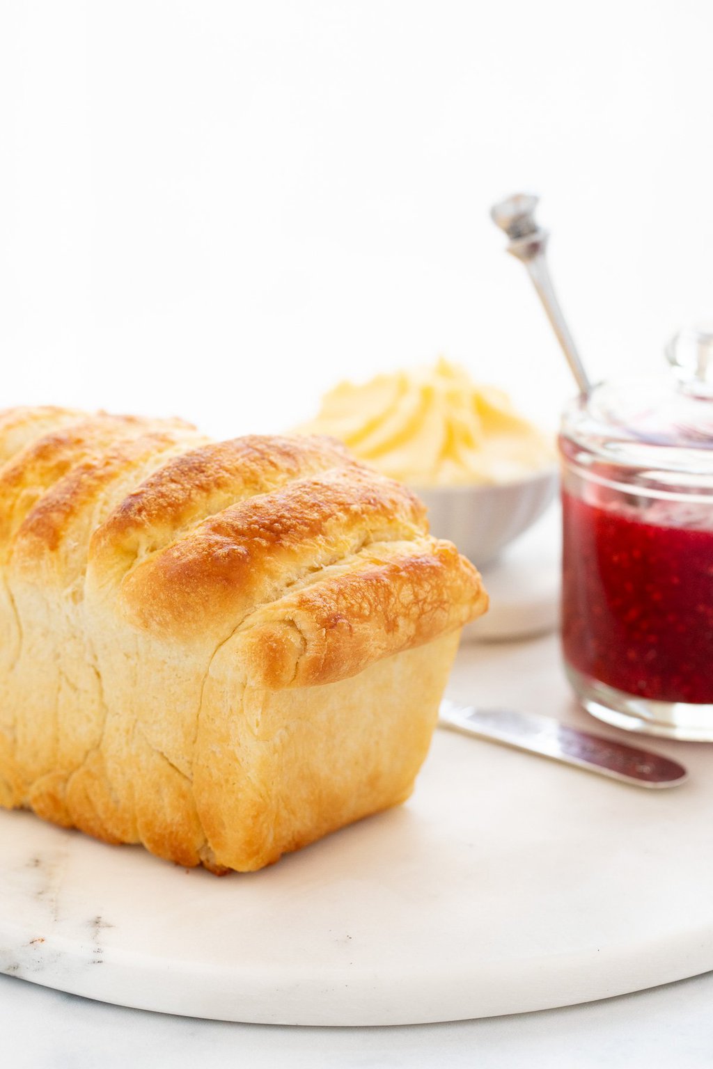 Vertical photo of a loaf of Pull Apart Brioche Bread on a round marble surface with a glass jar of raspberry jam and a dish of fresh butter in the background.