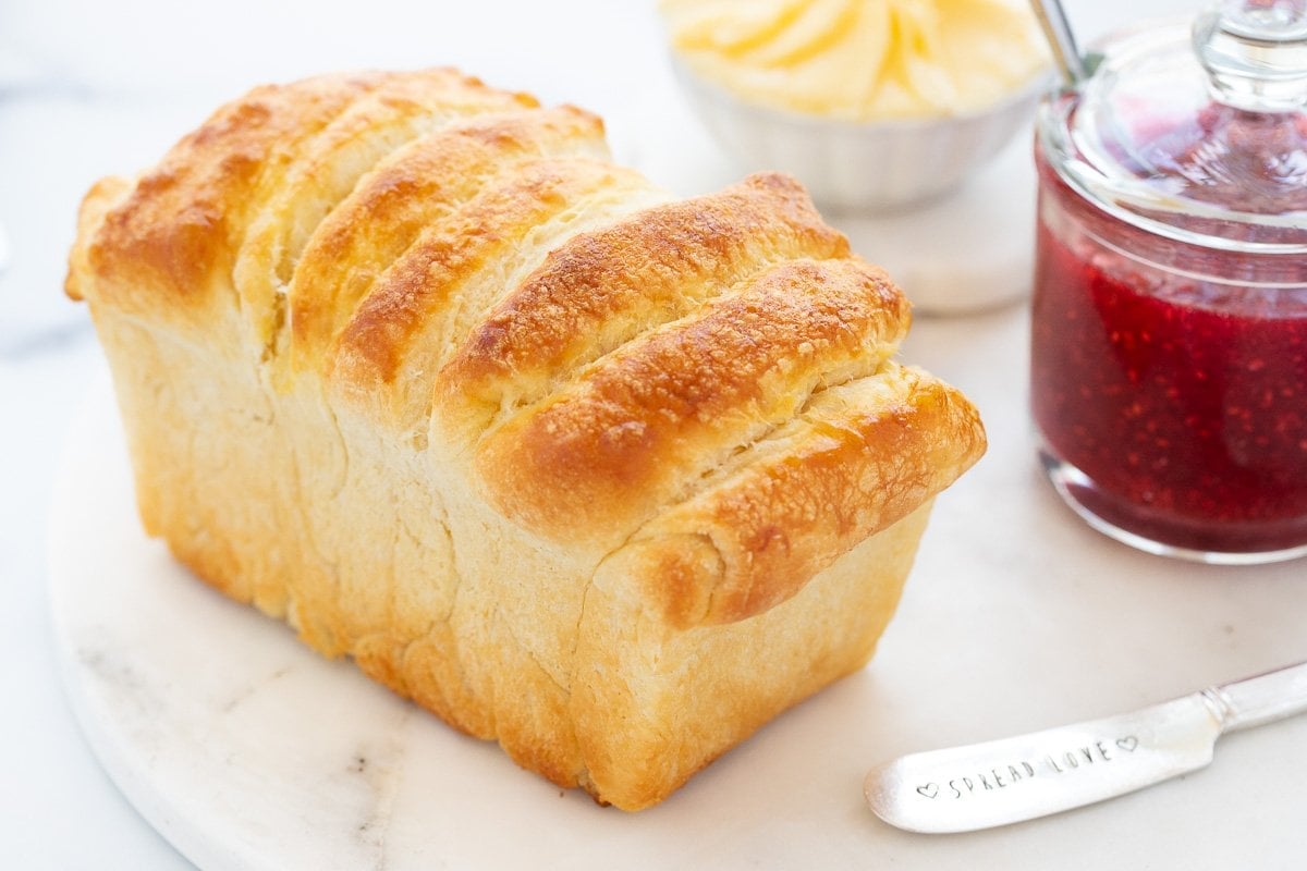 Horizontal closeup photo of a loaf of No Knead Pull-Apart Brioche Bread on a marble surface with a glass jar of raspberry jam and a dish of fresh butter in the background.