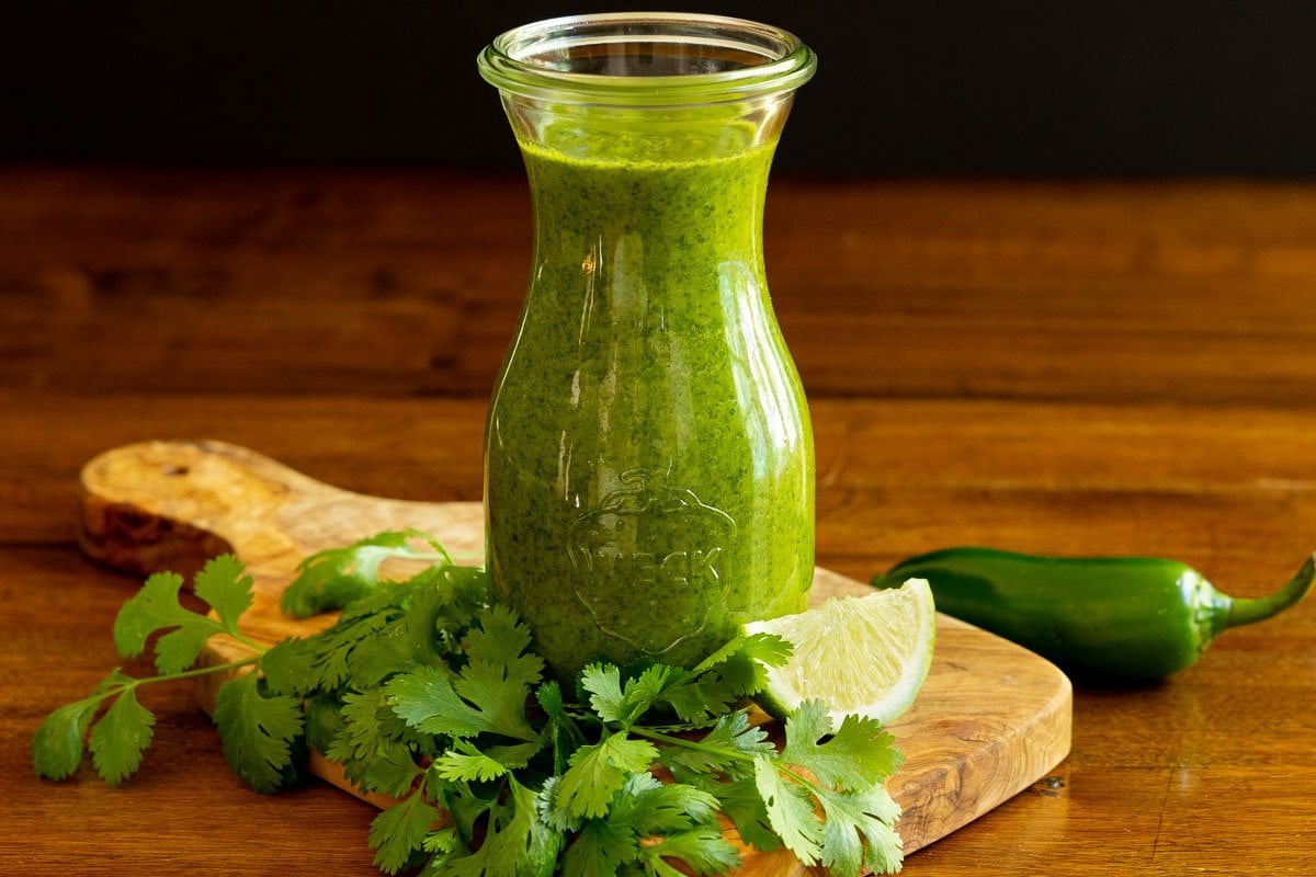 Horizontal photo of a Weck glass jar of Mexican Cilantro Sauce on a wood cutting board surrounded by fresh cilantro leaves and lime wedges.