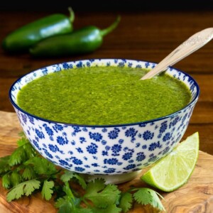 Vertical closeup photo of a serving bowl of Mexican Cilantro Sauce - thecafesucrefarine.com on a wood cutting board surrounded by lime wedges and fresh cilantro leaves.