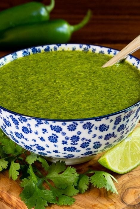 Horizontal photo of a blue and white patterned bowl filled with Mexican Cilantro Sauce surrounded by fresh cilantro leaves and lime wedges.