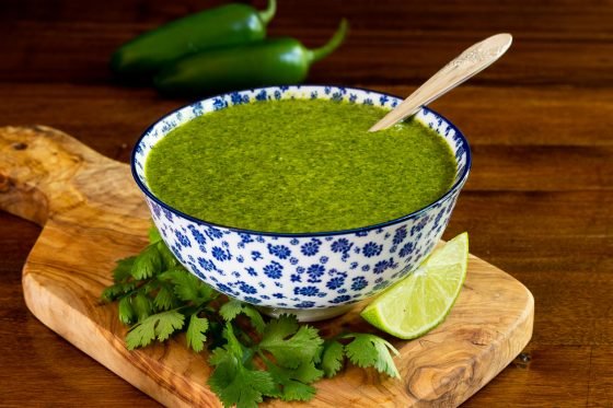Horizontal photo of a blue and white patterned bowl filled with Mexican Cilantro Sauce surrounded by fresh cilantro leaves and lime wedges.