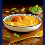 Vertical photo of a blue and white patterned serving bowl of Roasted Carrot Tomato Soup garnished with fresh basil leaves, roasted tomatoes and a zinnia flower.
