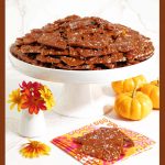 Vertical photo of Sea Salted Pumpkin Pecan Brittle on a white pedestal plate surrounded by mini pumpkins and zinnia flowers.