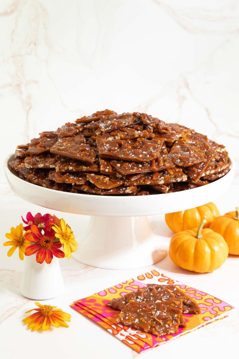 Vertical photo of Sea Salted Pumpkin Pecan Brittle on a white pedestal plate surrounded by mini pumpkins and orange zinnia flowers.