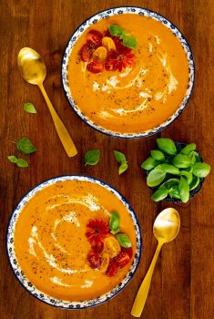Vertical overhead photo of two individual serving bowls of Roasted Carrot Tomato Soup on a wood table garnished with fresh basil leaves.