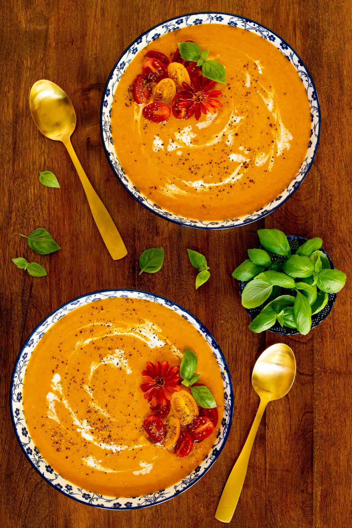 Vertical overhead photo of two serving bowls of Creamy Roasted Tomato Carrot Bisque on a wood table garnished with fresh basil leaves and a zinnia flower.