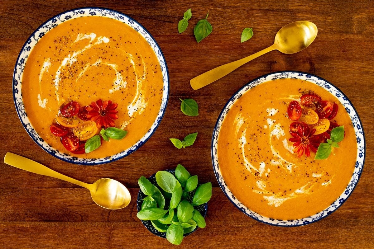 Horizontal overhead photo of two serving bowls of Roasted Carrot Tomato Soup on a wood table with garnishes of cream, sliced mini tomatoes and fresh basil.