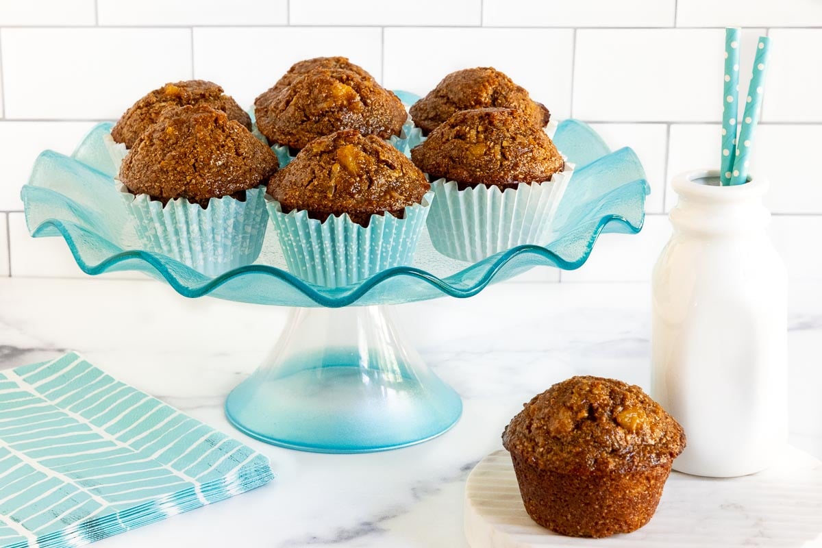 Horizontal photo of a batch of Honey-Glazed Pineapple Coconut Bran Muffins on a turquoise glass pedestal display plate.
