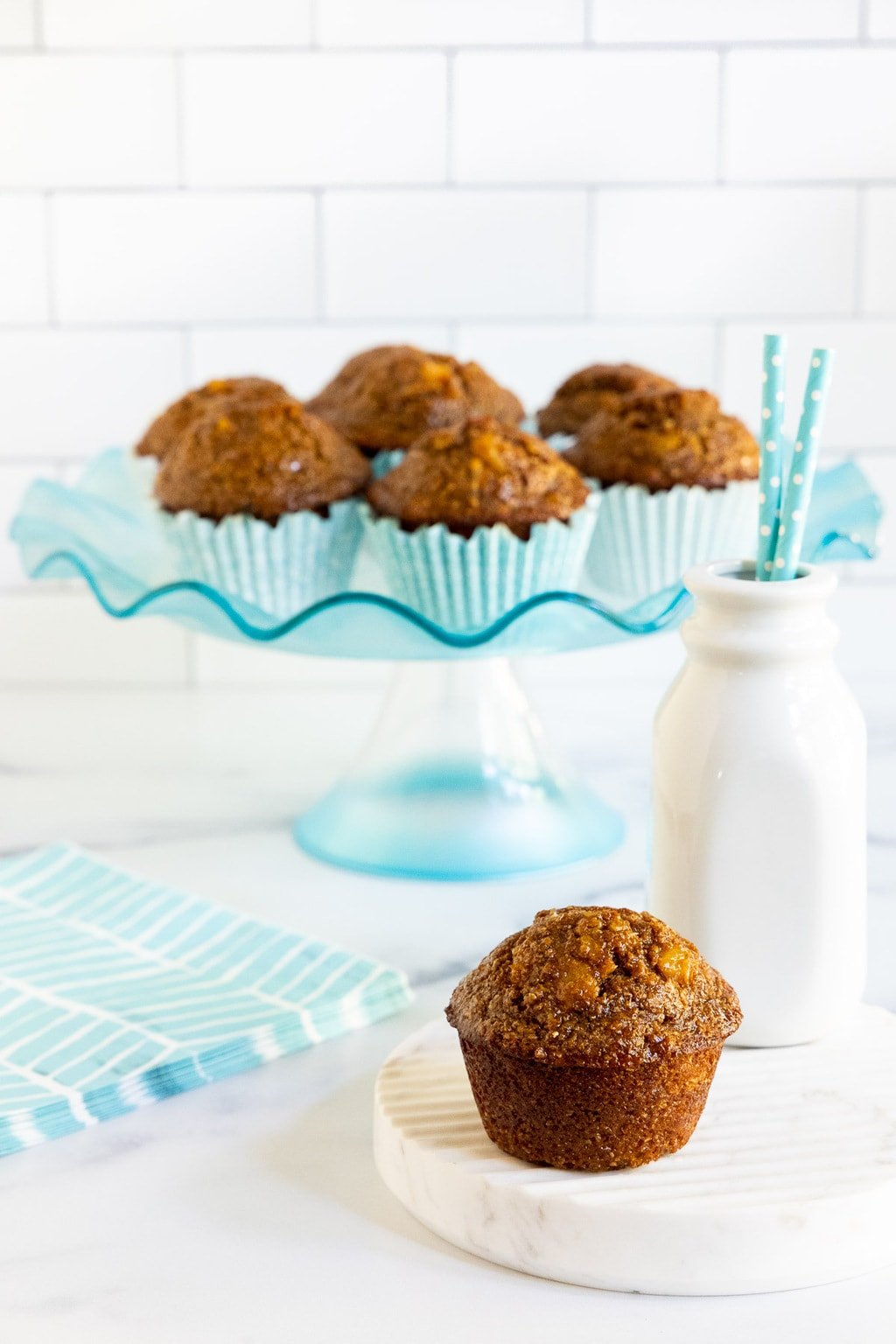 Vertical photo of a batch of Honey-Glazed Pineapple Coconut Bran Muffins on a turquoise glass pedestal platter.