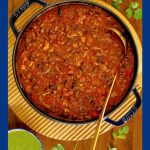 Vertical overhead photo of a Staub cast iron pot filled with Black Bean Turkey Chili and surrounded by lime wedges, pickled onions and cilantro sauce.