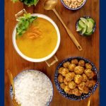 Vertical overhead photo of Thai Coconut Soup with Chicken Cilantro Meatballs surrounded by bowls of rice, lime wedges, fresh herb leaves and peanuts.