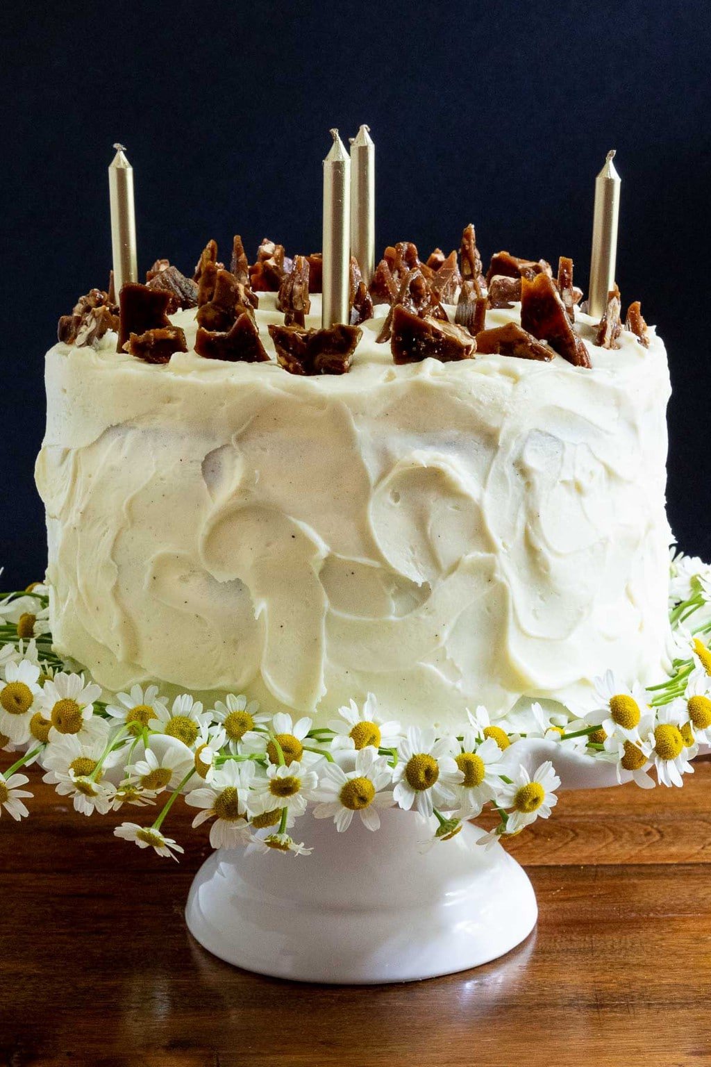Vertical photo of a Ridiculously Easy Applesauce Layer Cake on a white pedestal cake stand decorated with shards of Pumpkin Pecan Brittle and mini daisies.