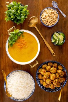 Vertical overhead photo of Thai Coconut Soup with Chicken Cilantro Meatballs on a wood table surrounded by bowls of peanuts, rice and fresh herbs.