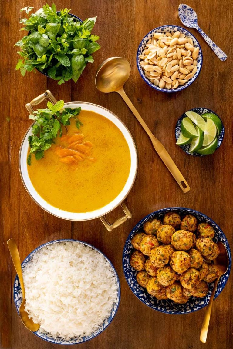 Vertical overhead photo of Thai Coconut Soup with Chicken Cilantro Meatballs on a wood table surrounded by bowls of peanuts, rice and fresh herbs.