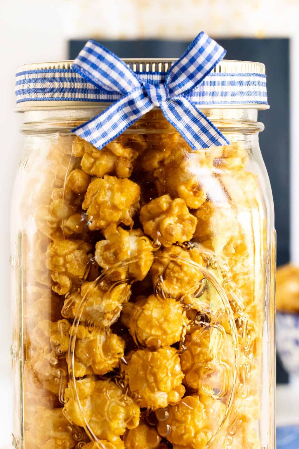 Vertical extreme closeup photo of a batch of Ridiculously Easy Sea Salted Microwave Caramel Corn in a wide mouth ball jar with a blue and white checkered bow for gift giving.