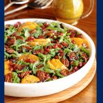 Vertical photo of a Cranberry Clementine Arugula Salad in a white marble serving bowl with a pitcher of salad dressing in the background.