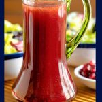 Vertical closeup photo of a glass cruet of Pomegranate Ginger Salad Dressing on a ribbed wood serving tray with two salads in the background.