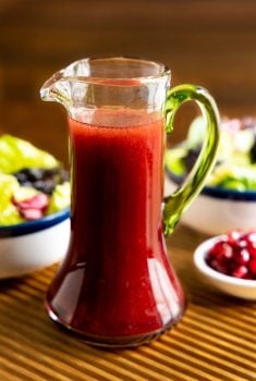 Vertical closeup photo of a cruet of Pomegranate Ginger Salad Dressing on a ribbed wood serving tray with fresh salads in the background.