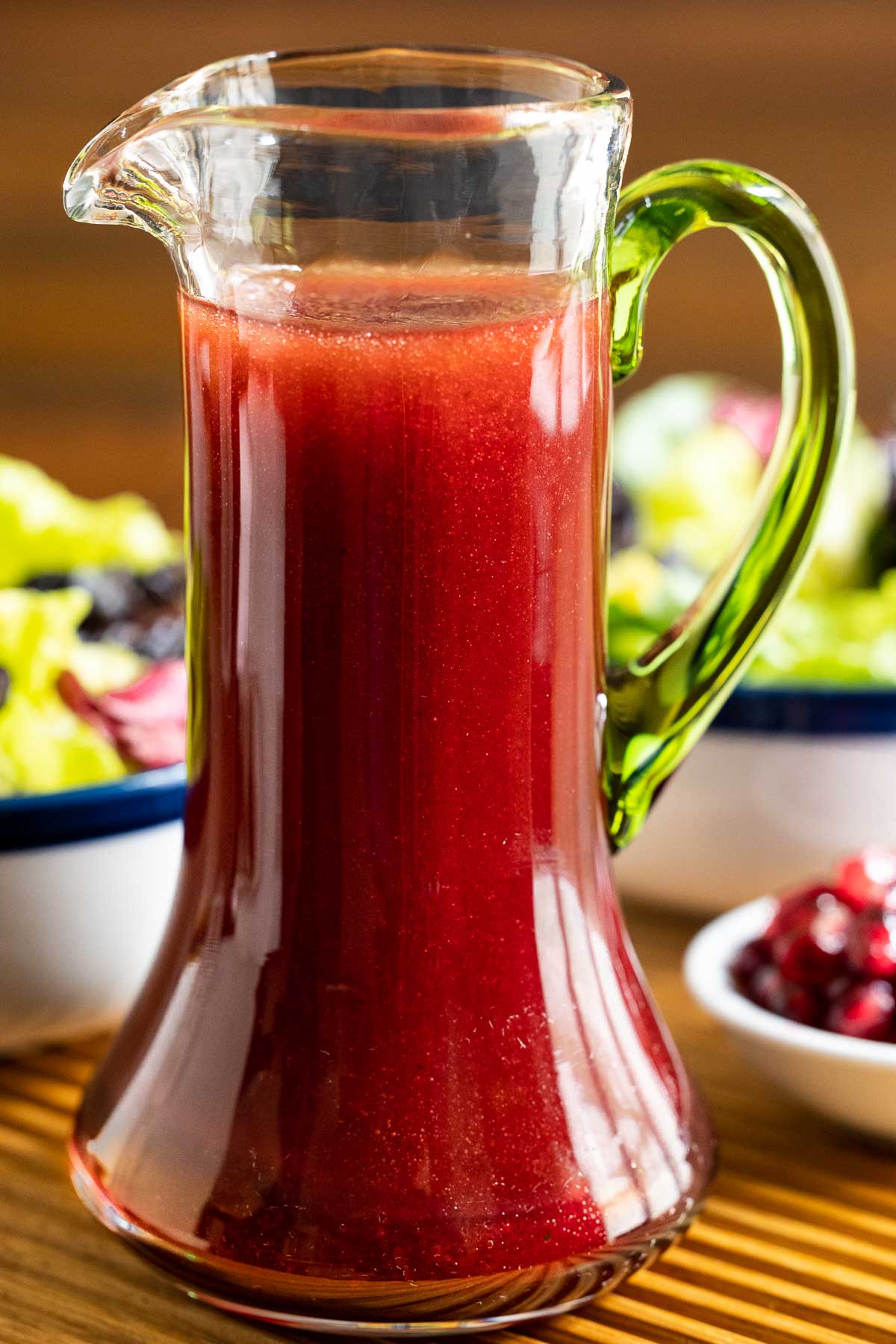 Vertical extreme closeup photo of a cruet of Pomegranate Ginger Salad Dressing on a ribbed wood tray with fresh salads in the background.