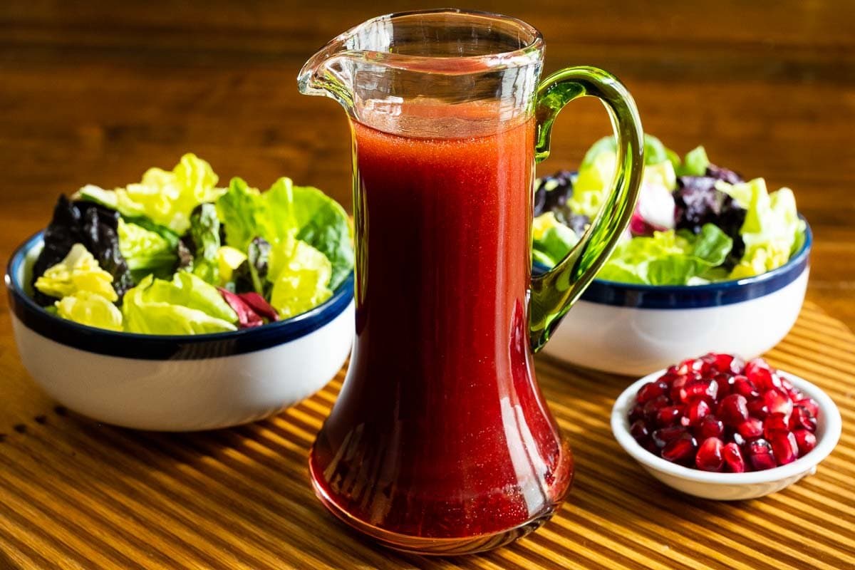 Horizontal photo of a cruet of Pomegranate Ginger Salad Dressing on a ribbed wood serving tray with two salads in the background.