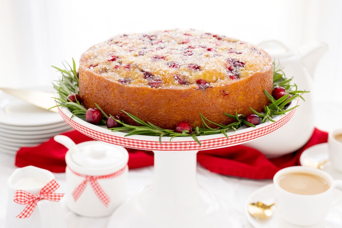 Horizontal photo of a Ridiculously Easy Orange-Glazed Cranberry Cake on a white pedestal cake stand decorated with red and white checkered ribbon.