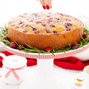 Horizontal photo of a Ridiculously Easy Orange-Glazed Cranberry Cake on a white pedestal cake stand.