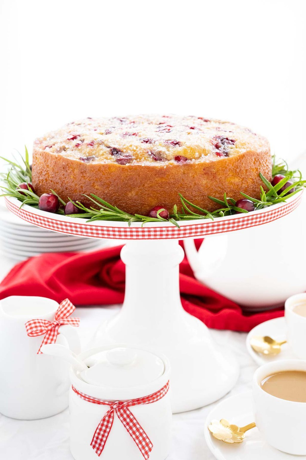 Vertical photo of a Ridiculously Easy Orange-Glazed Cranberry Cake on a white pedestal cake stand decorated with red and white checkered ribbon.