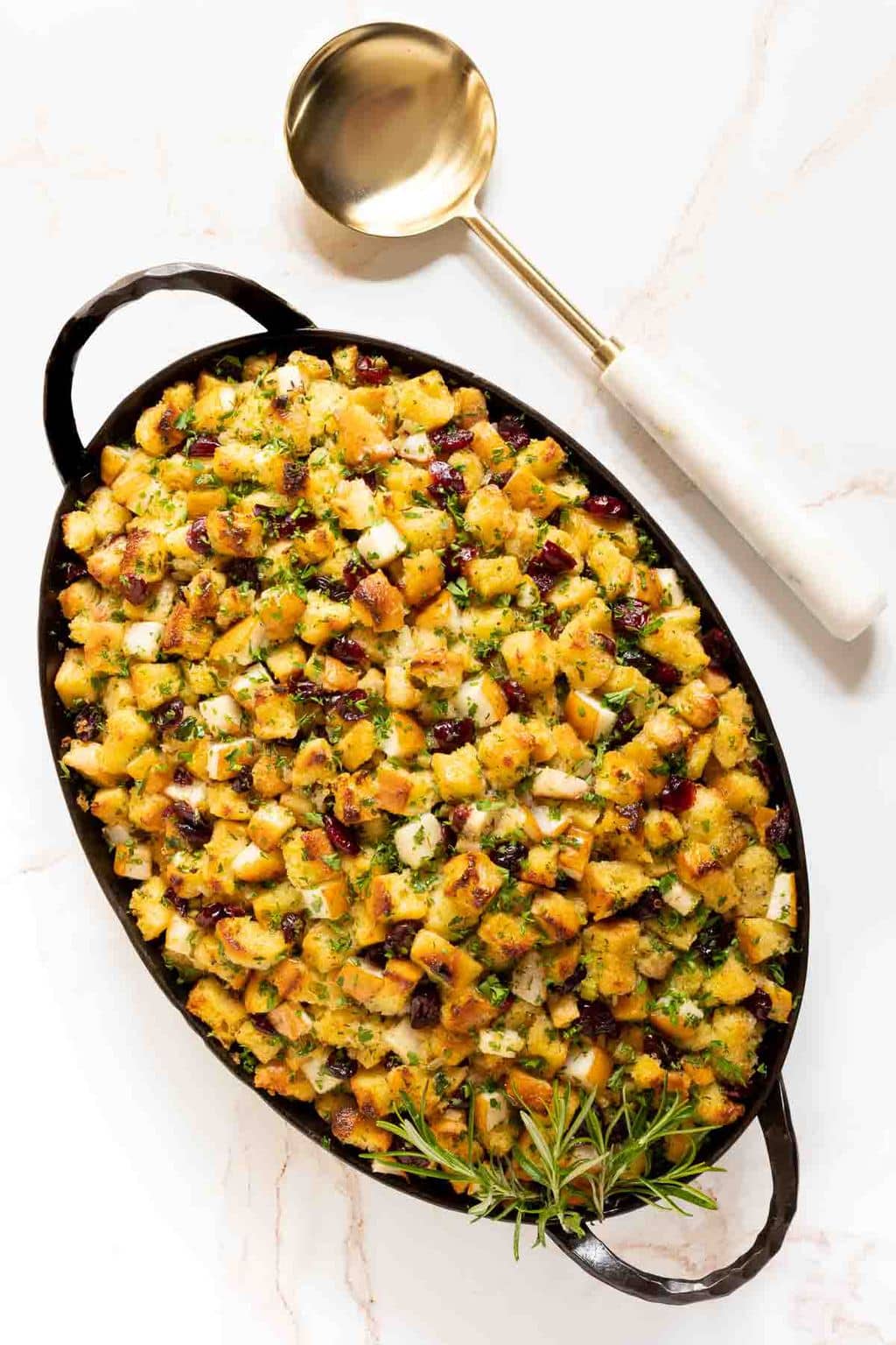 Vertical overhead photo of a hammered metal oval Smithey pan filled with The BEST Stuffing Recipe - Crispy, Buttery Herb Stuffing garnished with rosemary sprigs.