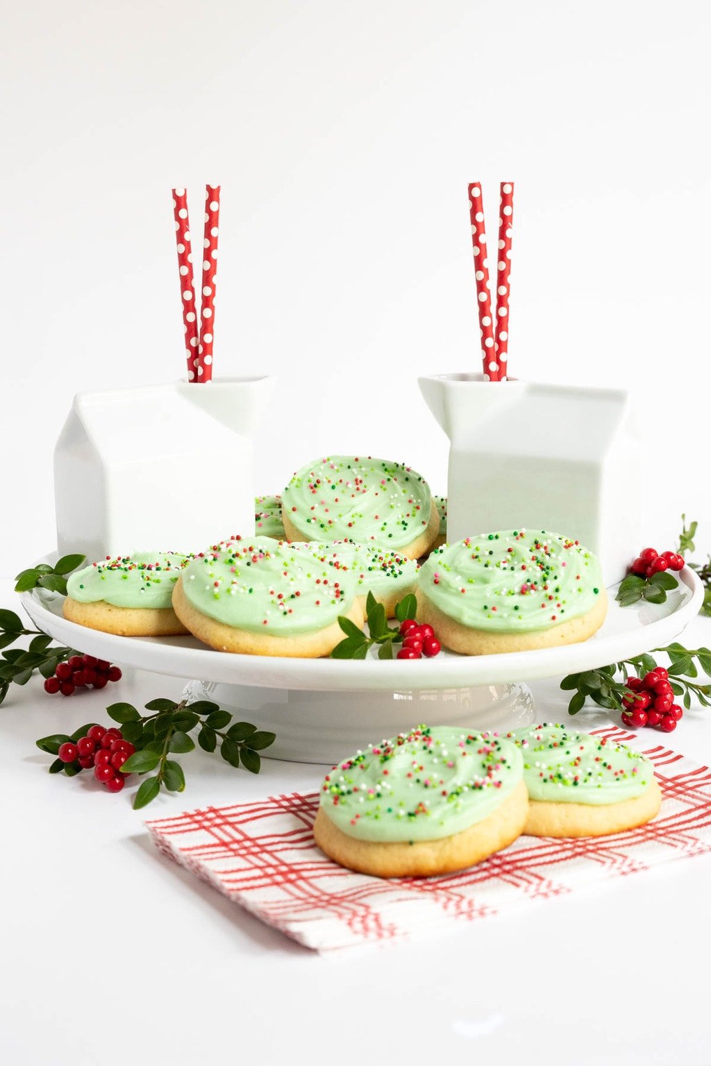 Vertical photo of Copycat Crumbl Christmas Sugar Cookies on a white pedestal serving plate surrounded by holly berries, cartons of milk and red and white napkins.