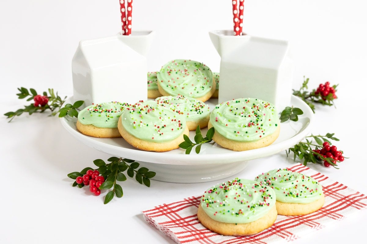 Horizontal photo of a batch of Copycat Crumbl Christmas Sugar Cookies on a white pedestal serving platter surrounded by milk containers and holly leaves and berries.