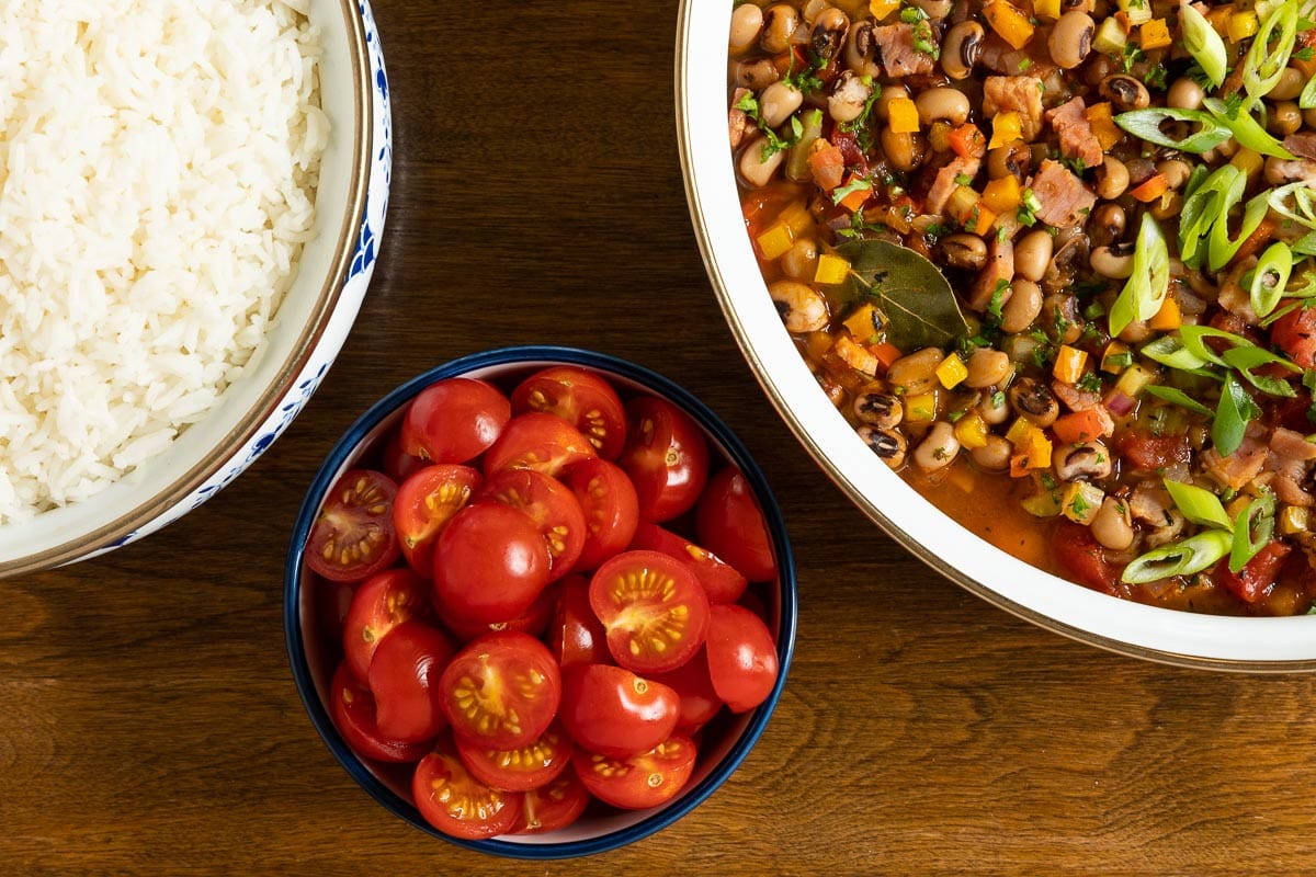 Horizontal overhead closeup photo of a pan of Healthy Hoppin' John next to a pot of white rice and a small bowl of sliced tomatoes.