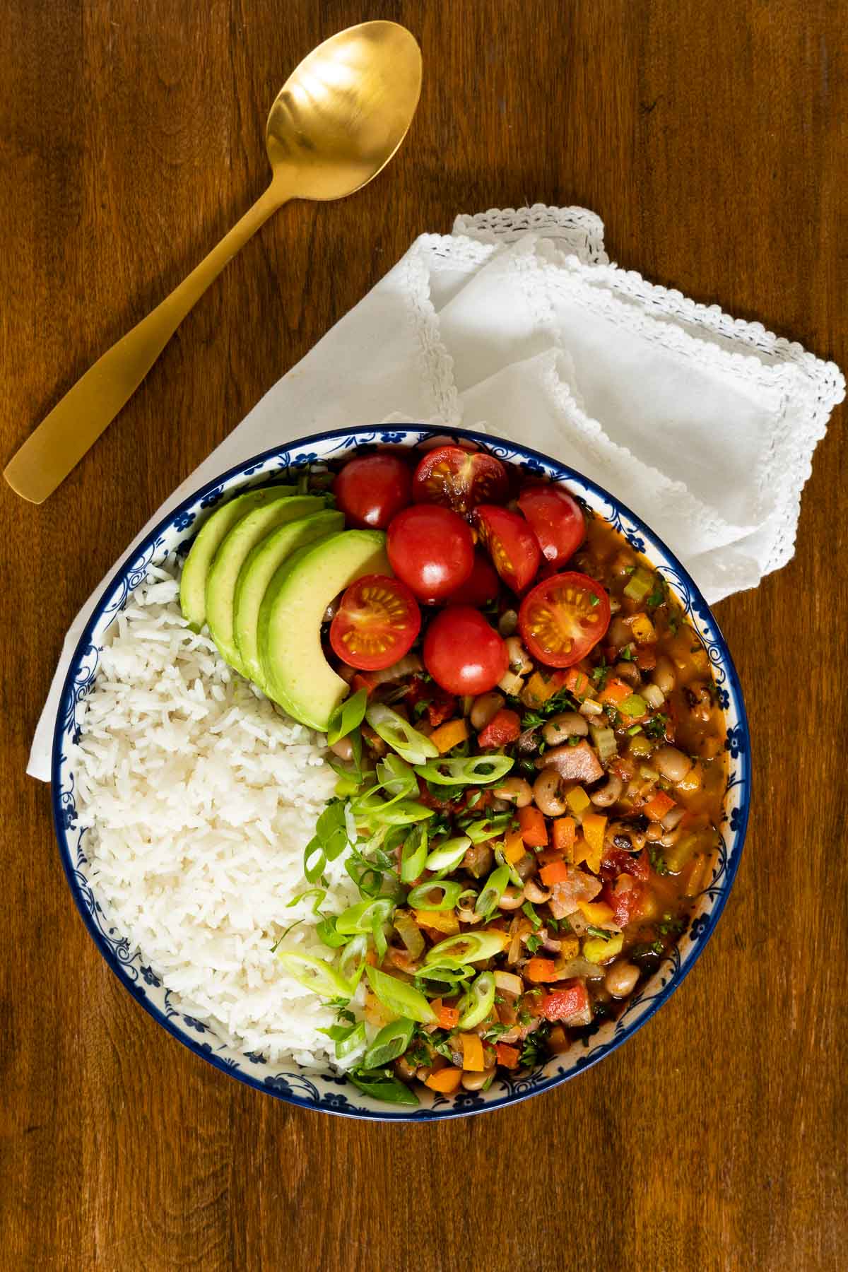 Vertical overhead photo of a serving bowl filled with Healthy Hoppin' John, a serving of white rice, sliced avocados, onions and tomatoes on a wood table.
