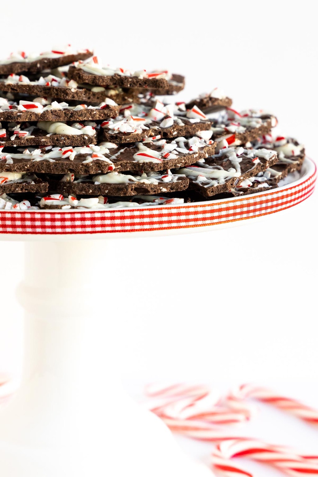 Vertical photo of a batch of Peppermint Bark Brittle on a white pedestal plate decorated with red and white checkered ribbon.