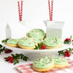 Vertical photo of a batch of Crumbl Christmas Sugar Cookies on a white pedestal serving platter surrounded by cartons of milk, holly berries and red and white patterned napkins.