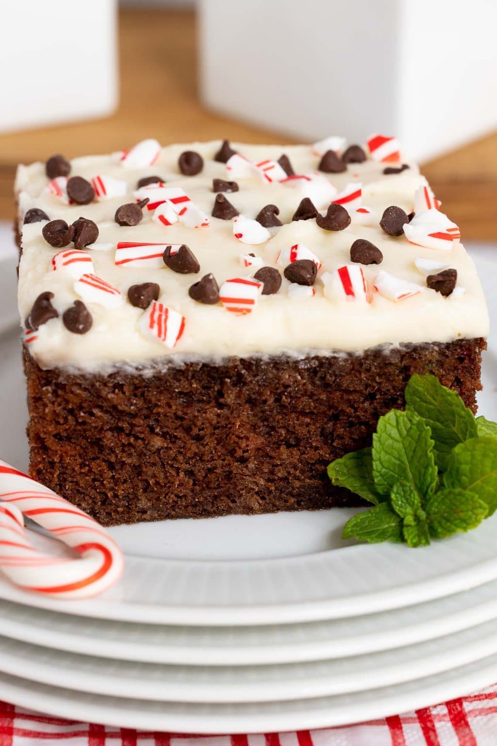 Vertical closeup photo of a slice of Ridiculously Easy Chocolate Peppermint Sheet Cake on a white serving plate surrounded by a candy cane and fresh mint leaves.