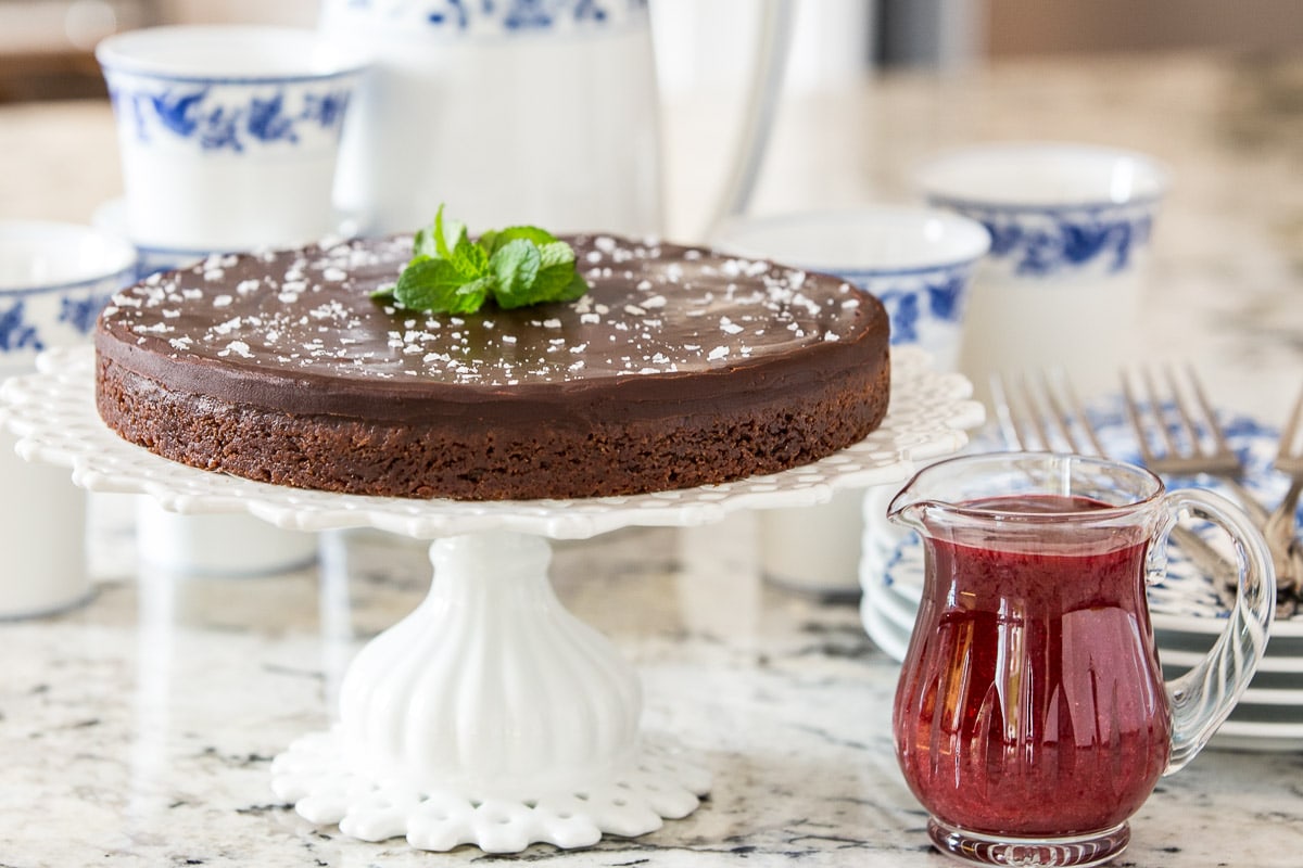 Horizontal photo of a Swedish Chocolate Sticky Cake (Kladdkaka) on a lattice pedestal serving plate with a blue and white milk pitcher and cup set in the background.