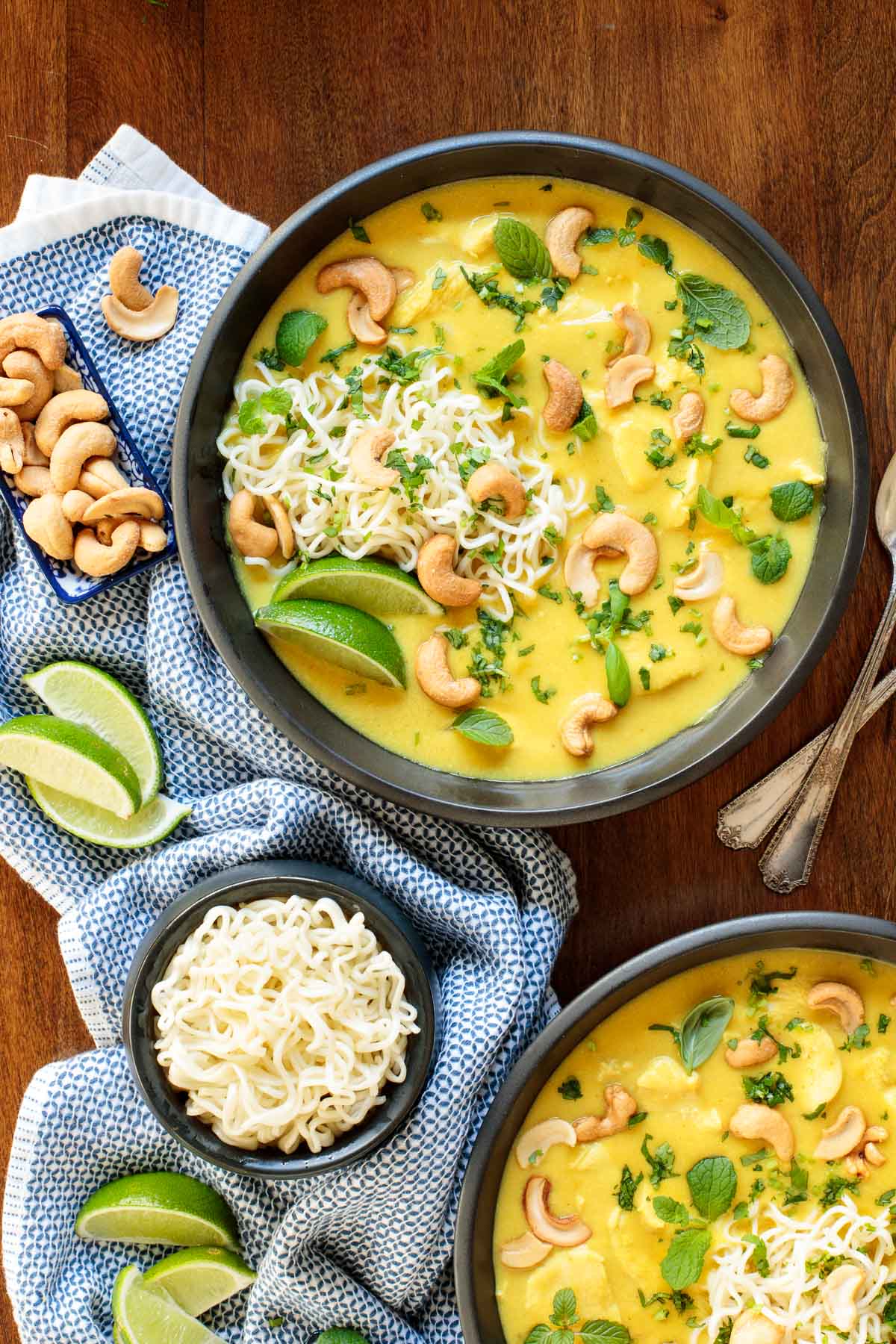Vertical overhead photo of two bowls of Coconut Curry Chicken Ramen on a wood table surrounded by a bowl of ramen noodles, lime wedges and cashews.
