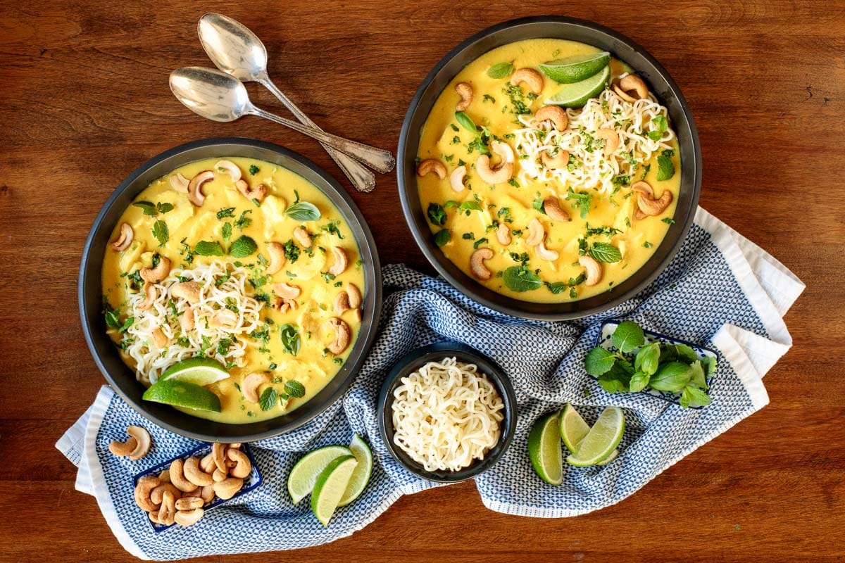 Horizontal overhead photo of two bowls of Coconut Curry Chicken Ramen on a wood table surrounded and garnished with cashews, ramen noodles, herbs and lime wedges.