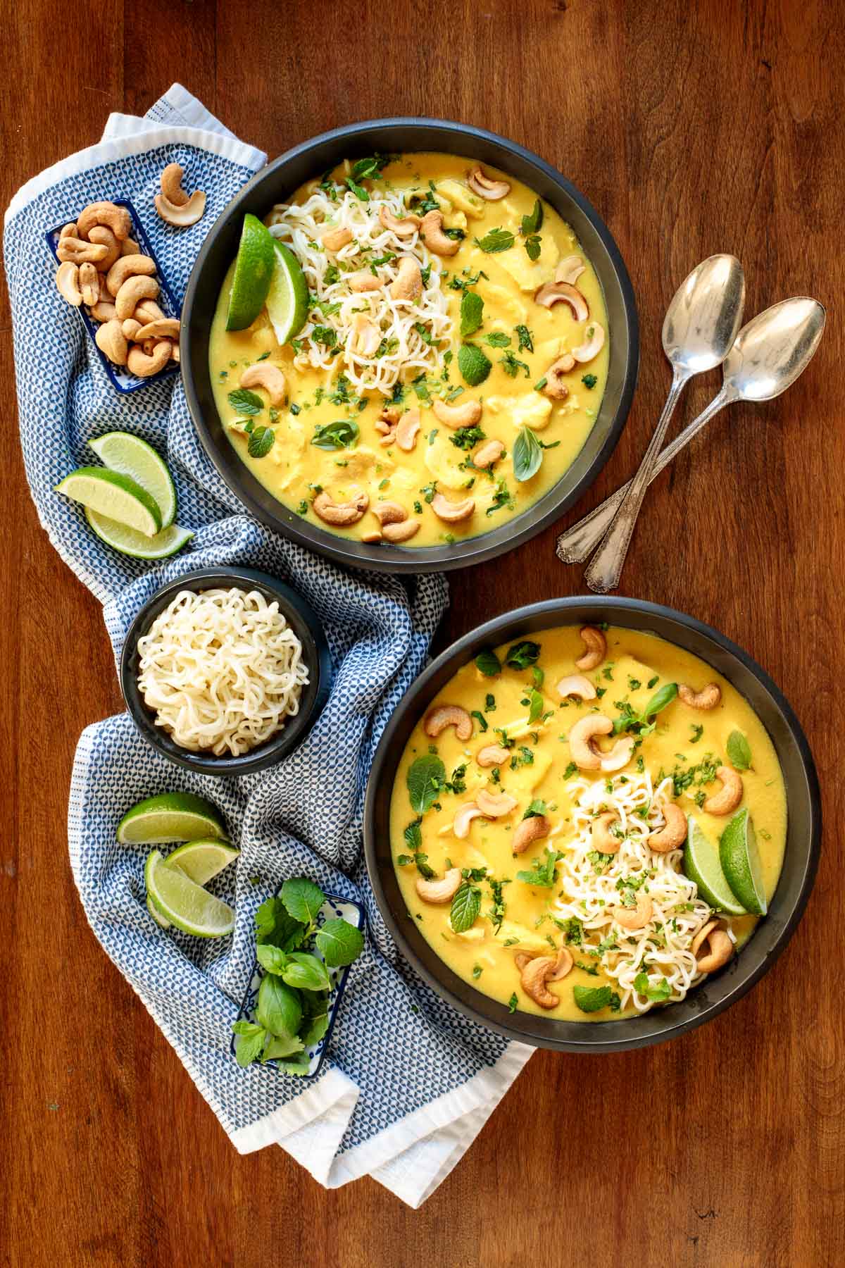 Vertical overhead photo of two black serving bowls of Coconut Curry Chicken Ramen on a wood table surrounded by lime wedges, fresh basil leaves, ramen and cashews.