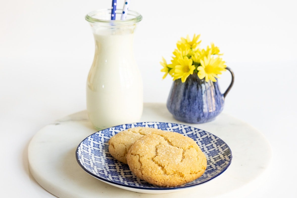 Horizontal photo of Crinkly Crackly Lemon Sugar Cookies on a blue and white patterned serving plate with a jar of milk and yellow flowers in the background.