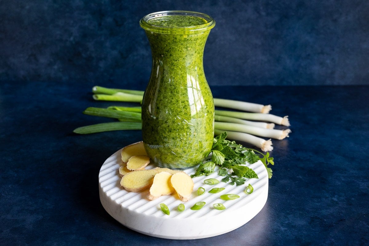 Horizontal photo of a cruet of Ginger Scallion Sauce on a granite trivet with fresh herbs and slices of ginger and scallions next to the bottle.