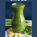 Vertical photo of a glass cruet of Ginger Scallion Sauce on a white granite trivet with slices of ginger and scallions along side fresh herbs. A title graphic is at the top of the photo.