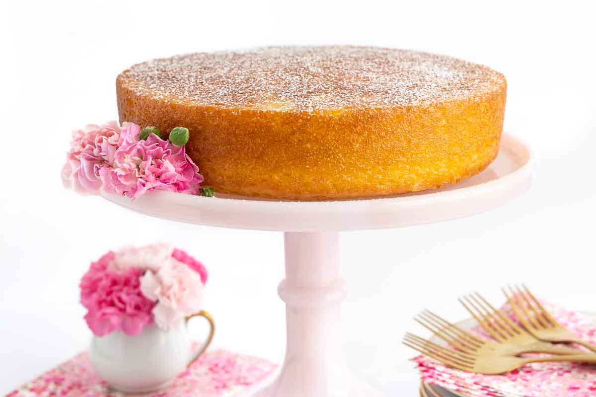 Horizontal photo of a Ridiculously Easy French Butter Cake on a pale pink pedestal cake stand decorated with pink flowers.