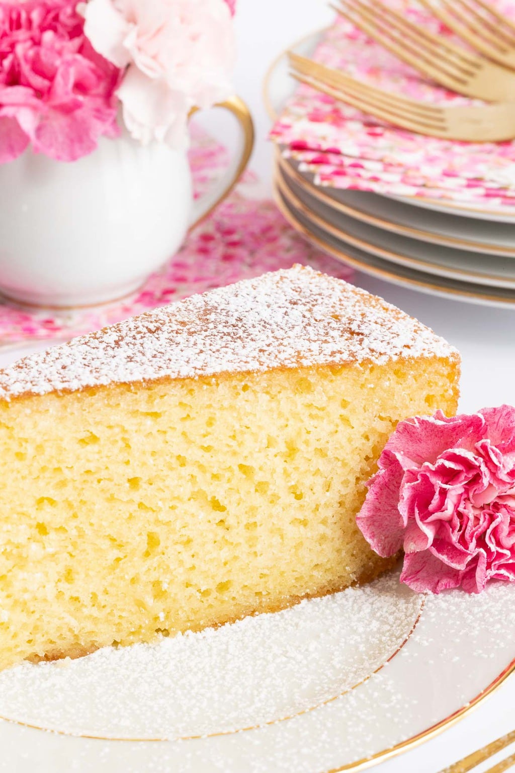 Vertical closeup photo of a slice of Ridiculously Easy French Butter Cake on a white and gold trimmed serving plate surrounded by pink carnation flowers.
