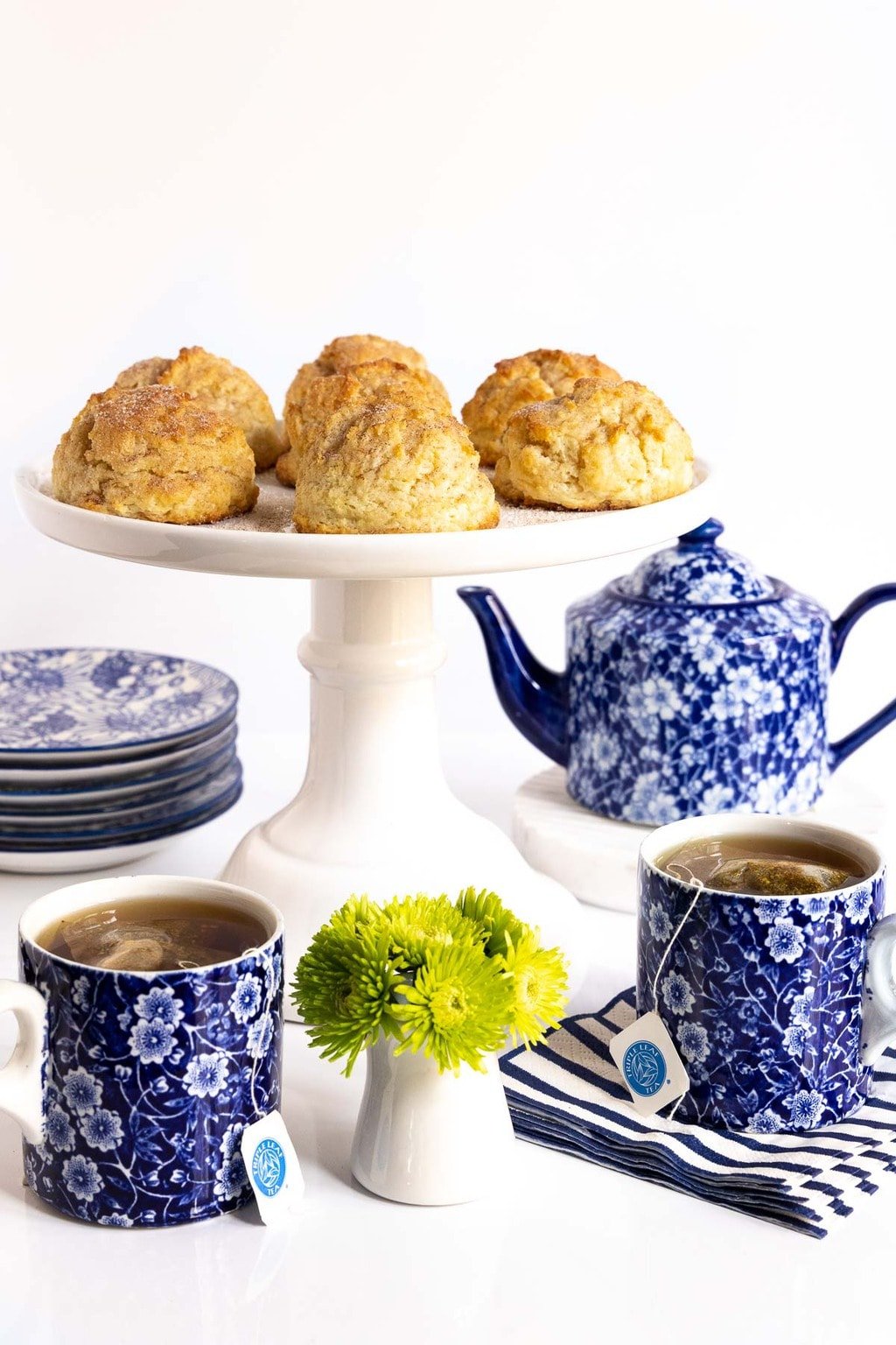 Vertical photo of a batch of Ridiculously Easy Snickerdoodle Scones on a white pedestal plate surrounded by a blue and white patterned tea set.