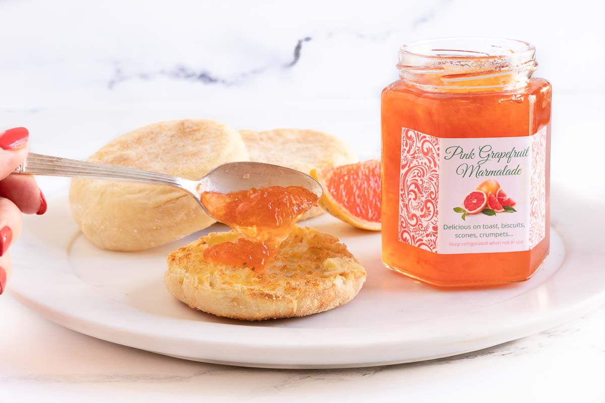 Horizontal photo of a jar of Pink Grapefruit Marmalade with a person spooning it on an English muffin.