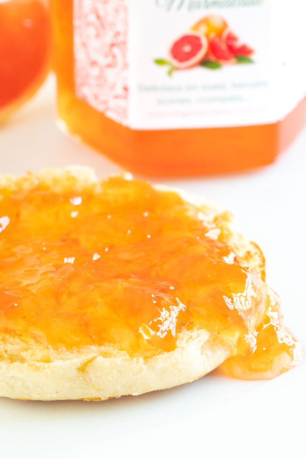 Vertical extreme closeup photo of an English muffin spread with Pink Grapefruit Marmalade and a jar of the marmalade in the background.