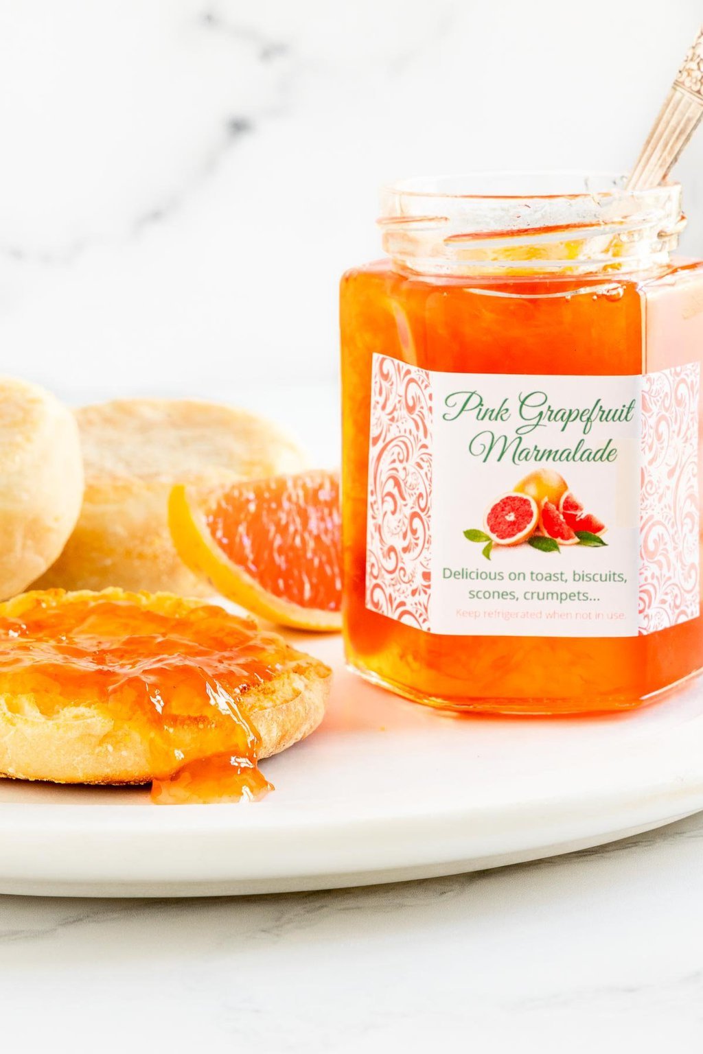 Vertical closeup photo of a jar of Pink Grapefruit Marmalade next to an English muffin.